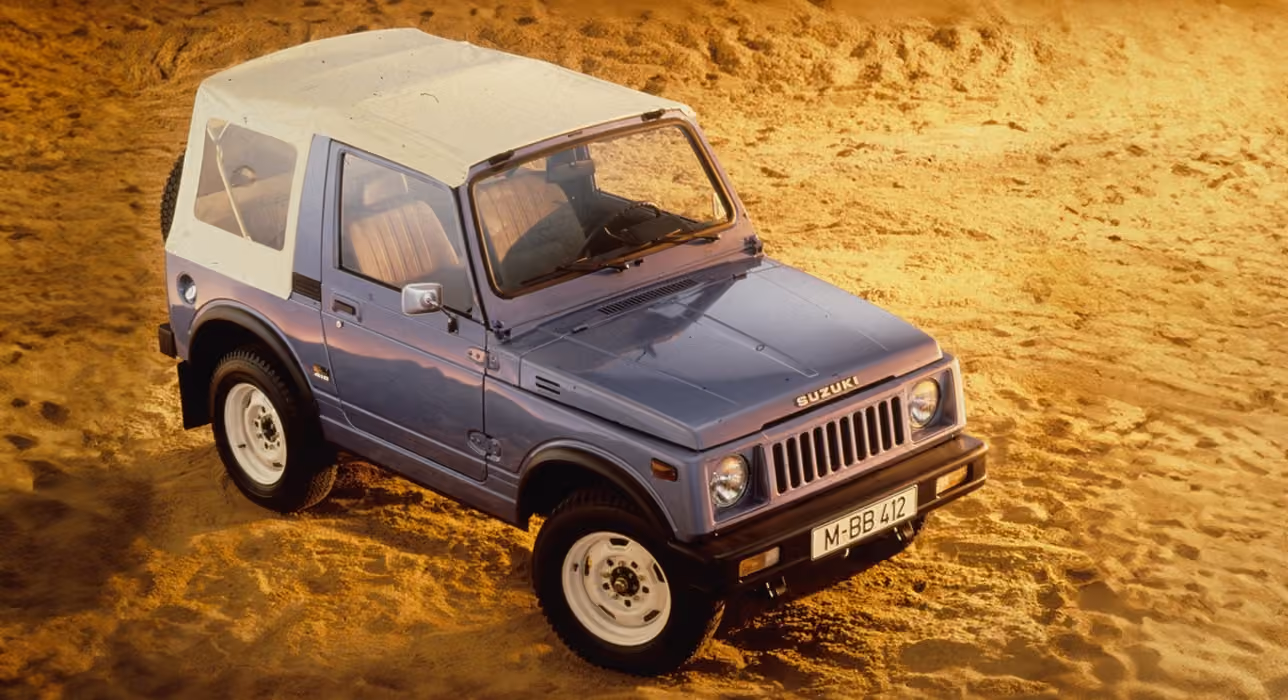 Abbildung zeigt von schräg oben einen blauen Jimny mit einem weißen Dach, der im Sand parkt