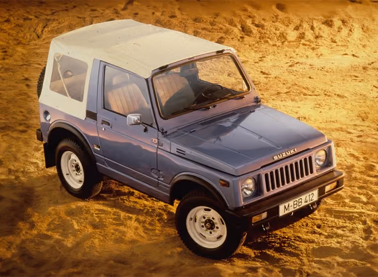 Abbildung zeigt von schräg oben einen blauen Jimny mit einem weißen Dach, der im Sand parkt
