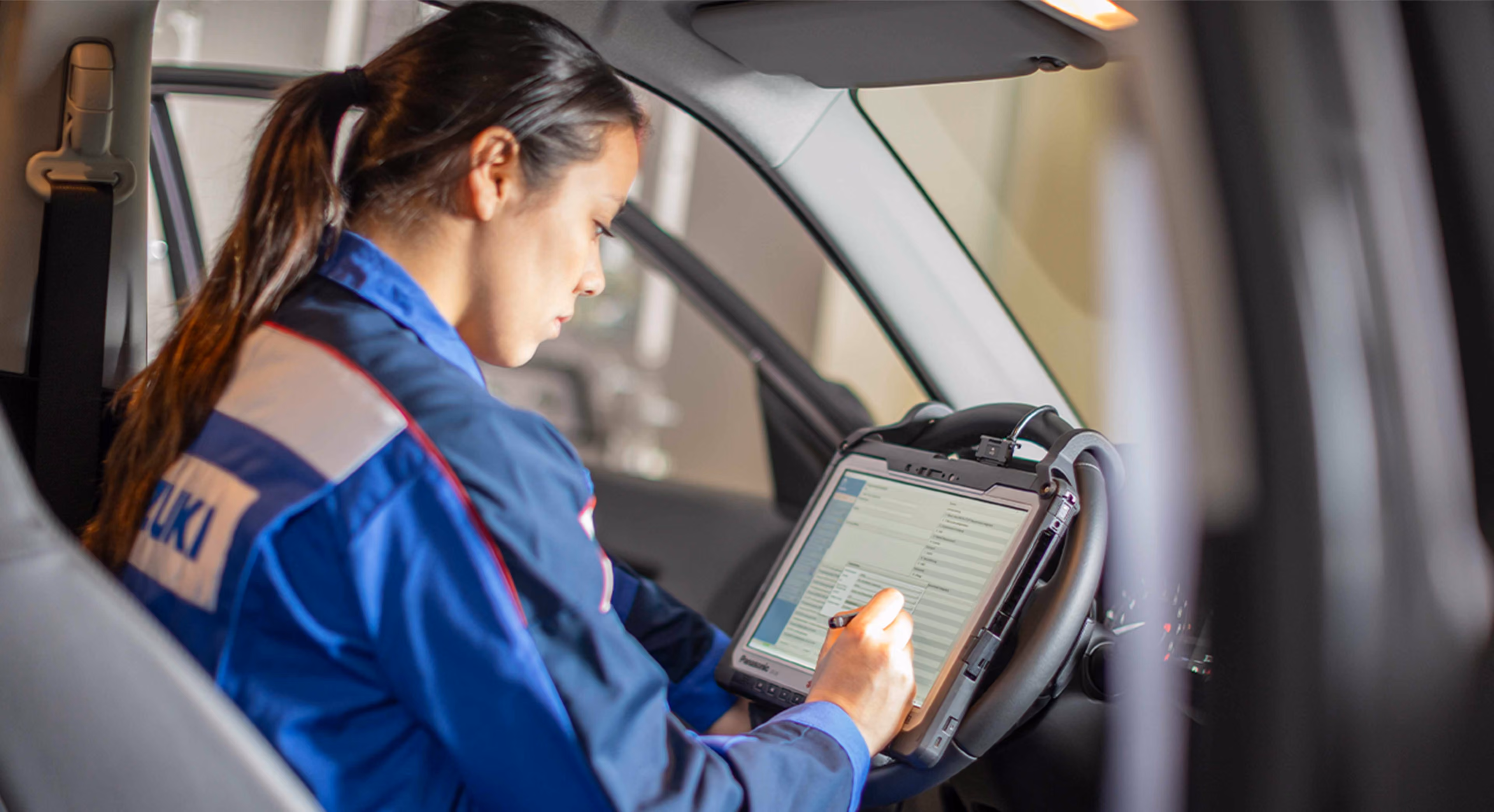 Eine Suzuki Mitarbeiterin sitzt in einem Suzuki Overall in einem Auto. Sie arbeitet an einer auf einem Tablett dargestellten Checkliste.