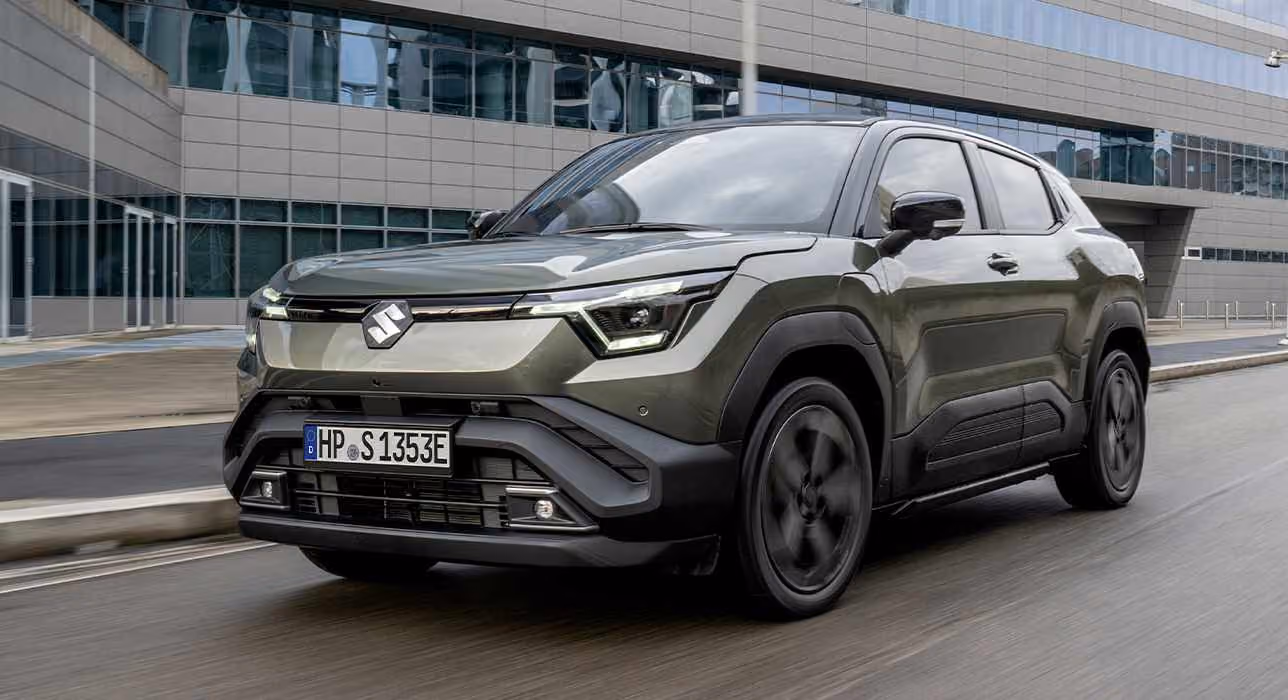 Ein dunkelgrüner e VITARA fährt auf einer Straße, im Hintergrund ist ein großes Gebäude mit Glasfront zu sehen.